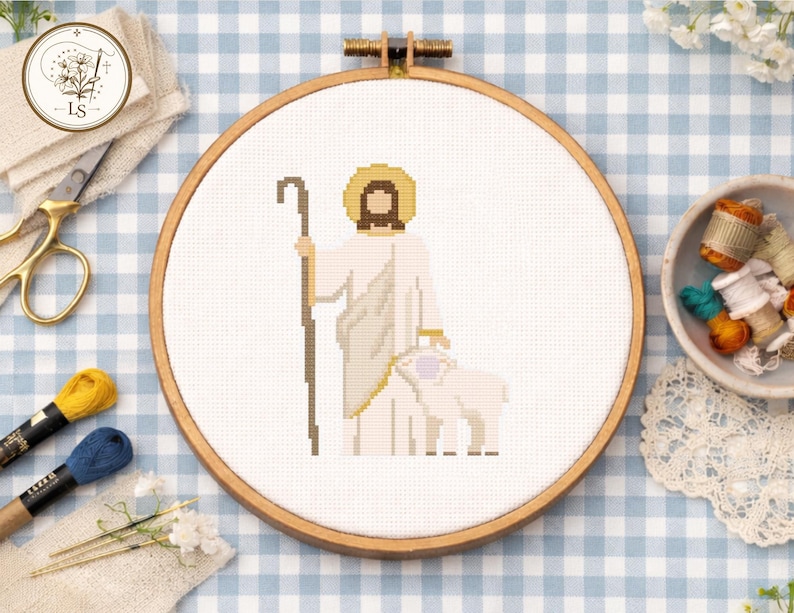 May include: A cross-stitch embroidery hoop featuring a religious figure with a staff, a halo, and a lamb. The design is in shades of white, beige, and brown, set against a white fabric. The wooden hoop is visible, and the background is a blue and white checkered pattern.