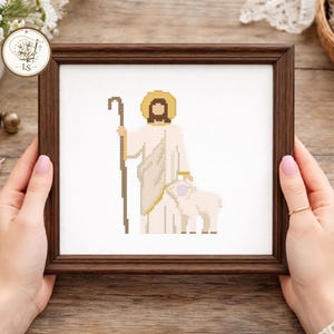 May include: A framed cross-stitch depicting a figure with a halo, holding a staff, and standing with a lamb. The artwork features a brown frame and is set against a white background. The figure is dressed in white and beige tones.