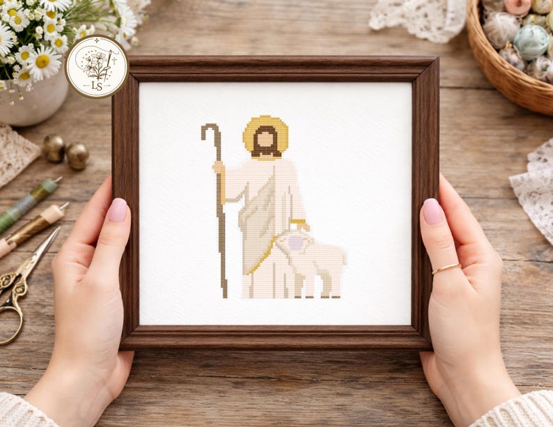 May include: A framed cross-stitch depicting a figure with a halo, holding a staff, and standing with a lamb. The artwork features a brown frame and is set against a white background. The figure is dressed in white and beige tones.