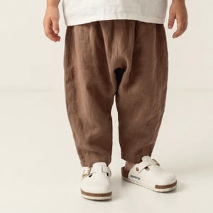 May include: Loose-fitting brown trousers with a tapered leg and an elasticized waistband, paired with white slip-on clogs featuring a side buckle and a cork-style sole. The trousers have a visible textured fabric finish and a relaxed silhouette.