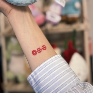 May include: A red temporary tattoo of three stacked circles on a person's forearm. The tattoo is located on the inner forearm, above the wrist. The person is wearing a blue and white striped button-down shirt.