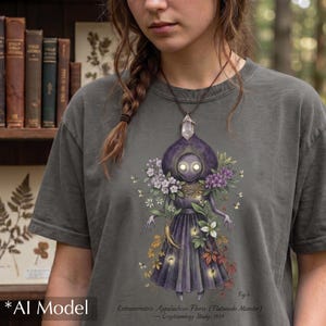 May include: A charcoal gray t-shirt featuring a graphic of a purple-robed figure with glowing eyes surrounded by flowers and foliage. The text reads: Fig. 6 Extraterrestris Appalachius Floris (Flatwoods Monster) — Cryptozoology Study, 1957.