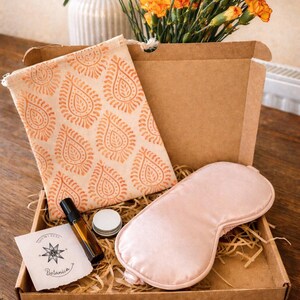 May include: A gift box containing a pink satin eye mask, a small essential oil bottle, a small jar, and a canvas pouch with an orange paisley pattern. The box also has a card with the word "Botanica". Yellow flowers are in a vase.