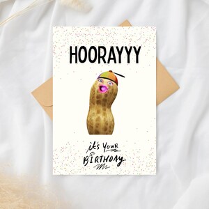 Peut inclure: Carte de vœux d'anniversaire avec le mot "HOORAYYY" en lettres noires grasses. La carte présente une cacahuète avec un visage portant un chapeau coloré. Le texte "it's your birthday" est écrit en dessous.