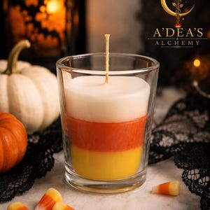 May include: A layered candle in a clear glass container featuring white, orange, and yellow wax stripes resembling candy corn. A single centered wick extends from the top. The text A'DEA'S ALCHEMY is printed in the upper right corner.