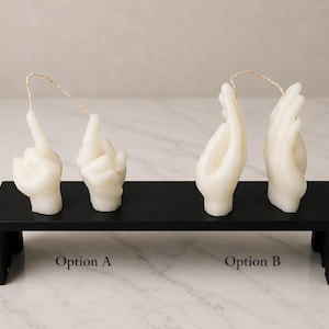 May include: Two sets of hand-shaped candles in ivory, each with a wick. The candles are displayed on a black rectangular stand. One set of candles is in the shape of a hand with the index finger pointing up, and the other set is in the shape of a hand with the fingers curled.