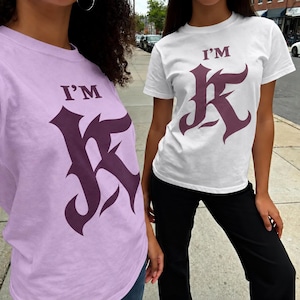 May include: Two short-sleeve t-shirts, one in light purple and one in white, each featuring the text "I'M" above a large, stylized letter "K" in a dark purple gothic-inspired font. The shirts have a crew neckline and a standard fit.