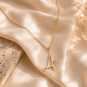 Pode incluir: Um colar de tom dourado com uma corrente fina e um pingente em forma de letra A cursiva, adornado com um pequeno detalhe de coração preso na parte superior direita da letra. O pingente está suspenso por um simples anel de ligação.