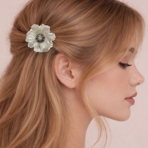 May include: A hair clip featuring a white, multi-petaled flower with a central circular embellishment. The flower has a pearlescent finish, while the center contains a small, round, light-colored stone surrounded by a ring of dark, metallic beads.