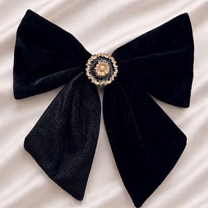 Könnte beinhalten: Eine schwarze Haarschleife aus Samt mit einer zentralen, kreisförmigen Verzierung. Das Schmuckelement zeigt in der Mitte ein goldenes Blumenmotiv, umgeben von einem dunklen, strukturierten Rand und einer gewellten, goldfarbenen Umrandung.
