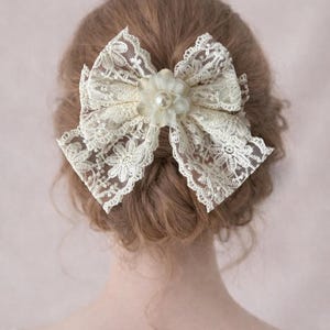May include: Ivory lace hair bow with a delicate floral center. The bow is made of intricate lace and is attached to a hair clip. The floral center features a pearl accent. This hair accessory adds a touch of vintage charm.