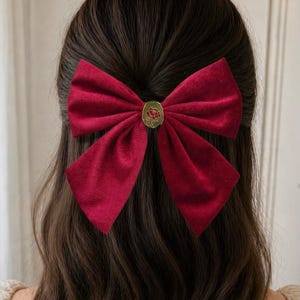 Könnte beinhalten: Burgunderfarbene Haarspange mit Samtschleife und goldfarbenem Blumendetail. Die Schleife sitzt in dunkelbraunem Haar und bildet einen auffälligen Kontrast. Das Haaraccessoire verleiht Eleganz.