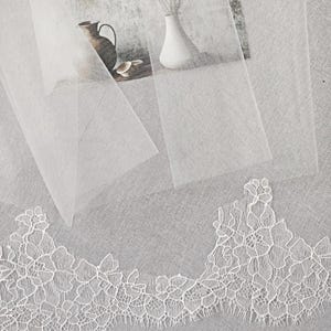 Puede incluir: Primer plano de un ribete de encaje blanco con un delicado dise&ntilde;o floral. El encaje se coloca sobre una tela blanca transparente, con un fondo suave y texturizado. Tambi&eacute;n se ve un jarr&oacute;n con flores blancas y una jarra decorativa.