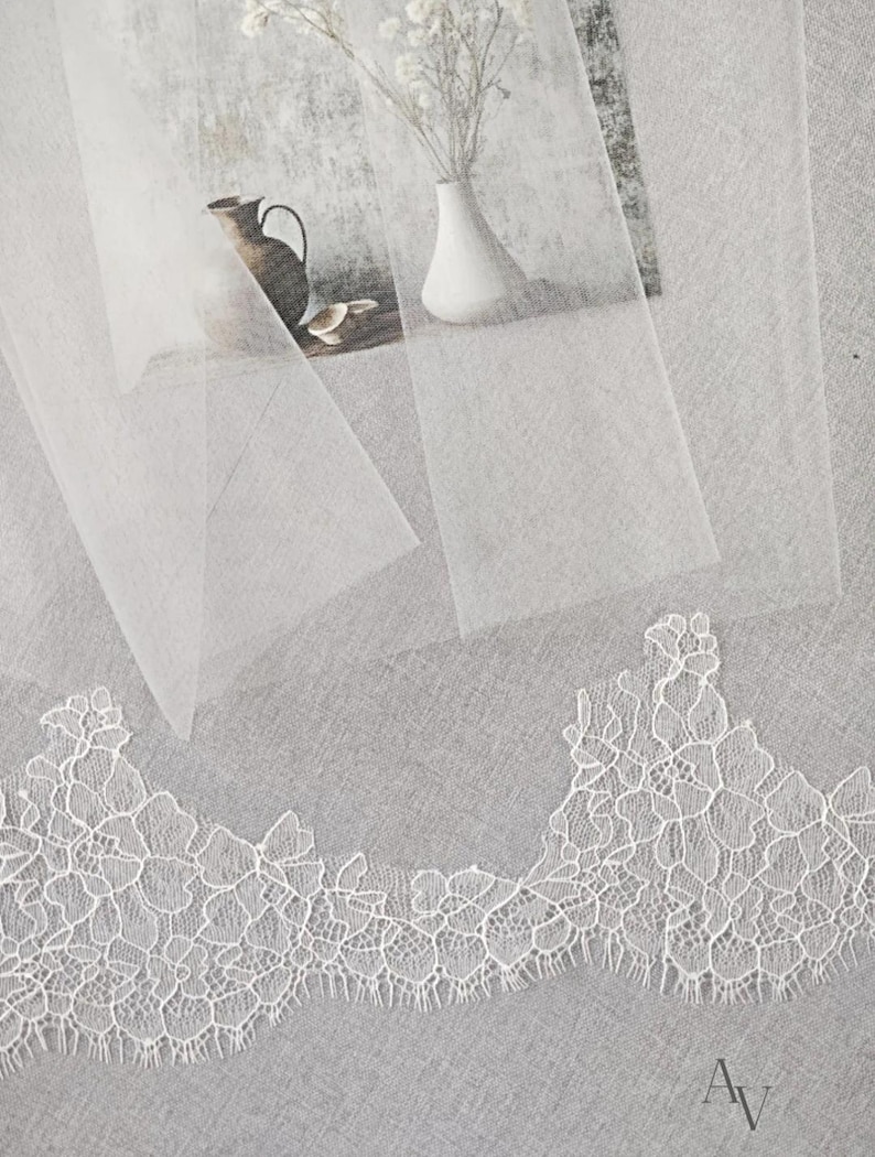 Puede incluir: Primer plano de un ribete de encaje blanco con un delicado dise&ntilde;o floral. El encaje se coloca sobre una tela blanca transparente, con un fondo suave y texturizado. Tambi&eacute;n se ve un jarr&oacute;n con flores blancas y una jarra decorativa.