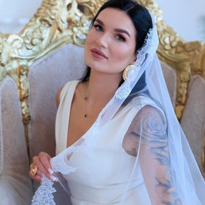 Puede incluir: Una novia con un vestido blanco sin mangas y un velo transparente con delicados bordes de encaje. Est&aacute; sentada en una silla dorada ornamentada y lleva pendientes y un anillo dorados. El velo est&aacute; levantado, revelando un tatuaje en su brazo.