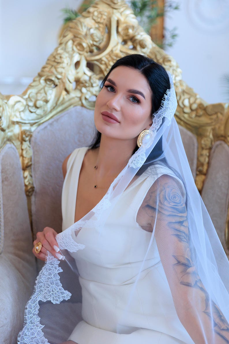 Puede incluir: Una novia con un vestido blanco sin mangas y un velo transparente con delicados bordes de encaje. Est&aacute; sentada en una silla dorada ornamentada y lleva pendientes y un anillo dorados. El velo est&aacute; levantado, revelando un tatuaje en su brazo.