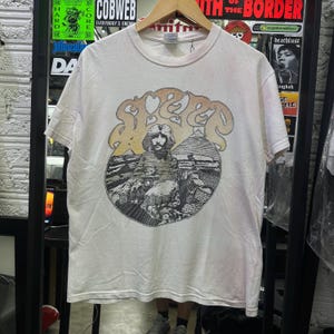 Könnte beinhalten: Ein cremefarbenes Kurzarm-T-Shirt mit einem Grafikdruck des Wortes SLEEP in einer stilisierten Schriftart über einer Illustration einer Sphinx und Pyramiden. Das Design ist in Schwarz-, Grau- und gedämpften Gelbtönen auf der Brust gehalten.