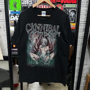 May include: Black t-shirt featuring the band name "Cannibal Corpse" in a distressed, green font above a graphic. The graphic depicts a nude figure surrounded by vines and the text "Worm Infested" in green. The shirt is hanging on a wooden hanger.
