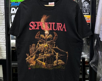Puede incluir: Una camiseta negra de manga corta con la palabra SEPULTURA en letras rojas sobre una ilustración gráfica detallada en tonos naranja, amarillo y marrón. La obra representa una escena oscura y surrealista con un ojo central y estructuras de piedra.