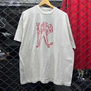 May include: A cream-colored short-sleeve t-shirt featuring a red graphic print of a devil figure holding a trident with the number 13 on its chest. The shirt has a crew neckline and is displayed on a wooden hanger against a chain-link fence.