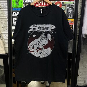 May include: A black short-sleeve t-shirt featuring a white graphic print of a figure riding a winged creature on a rocky surface against a dark red circular background. The word SLEEP is printed in a stylised white font above the graphic.