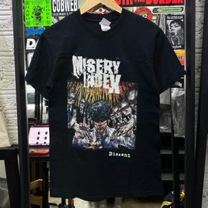 Puede incluir: Una camiseta negra de manga corta con un estampado gráfico que incluye el texto Misery Index en la parte superior y Dissent en la inferior. La ilustración muestra a una figura central con camisa blanca rodeada de personas con traje y gafas.