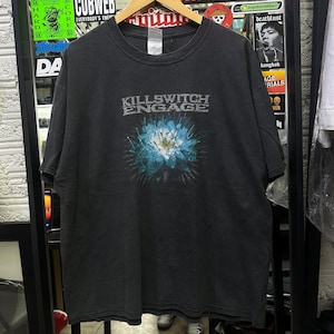 May include: A charcoal grey short-sleeve t-shirt featuring the text KILLSWITCH ENGAGE in a distressed white font above a graphic of a blue lotus flower with sharp, needle-like rays radiating outward from the centre of the blossom.