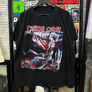 Puede incluir: Una camiseta negra de manga corta con un estampado gráfico en la parte delantera. El texto "CANNIBAL CORPSE" aparece en una fuente roja que gotea sobre una ilustración de una figura pálida y esquelética con acentos rojos y oscuros.