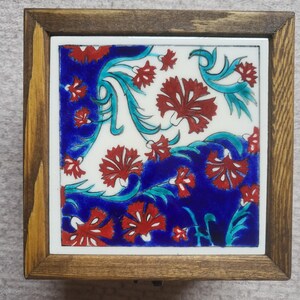 May include: A square wooden box featuring a decorative ceramic tile inlay on the lid. The tile displays a vibrant pattern of red carnation flowers and teal green leaves set against a deep blue and white background, framed by a dark wood border.