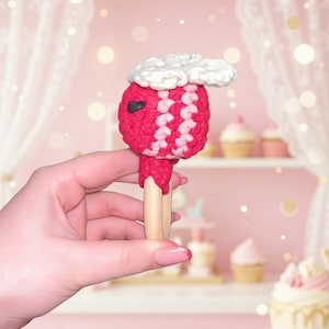 May include: A handmade, red and white crocheted lollipop with a white cloud-shaped top. The lollipop has a wooden stick and a small black heart-shaped detail. The background is a soft pink with decorative elements.