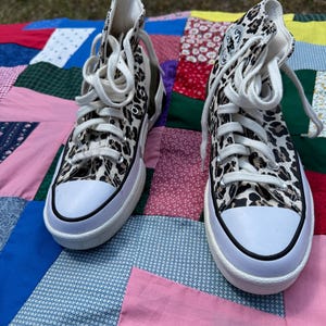 May include: Pair of high-top sneakers with a leopard print pattern. The shoes have white laces, a white toe cap, and a thick white sole with a black stripe. The background is a colorful patchwork quilt.