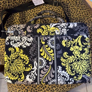 May include: A quilted laptop bag featuring a black, white, and yellow floral paisley pattern. The bag includes two top handles, a detachable black shoulder strap with metal hardware, and a rectangular shape with a zippered closure and vertical trim.