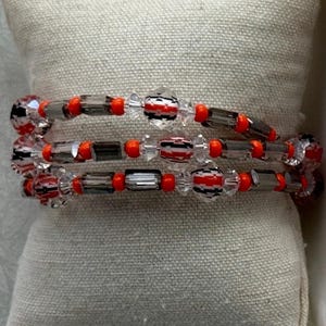 Puede incluir: Una pulsera de varias vueltas con un patrón repetitivo de cuentas redondas de color naranja brillante, cuentas de cristal transparente facetado y cuentas rectangulares de color gris oscuro, incluyendo cuentas esféricas con rayas negras, blancas y rojas.