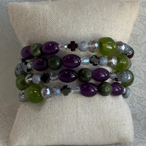 May include: A multi-strand wrap bracelet featuring a mix of smooth purple oval beads, dark green round beads, faceted clear crystals, grey marbled spheres, and small black four-leaf clover-shaped spacers arranged in a repeating pattern.