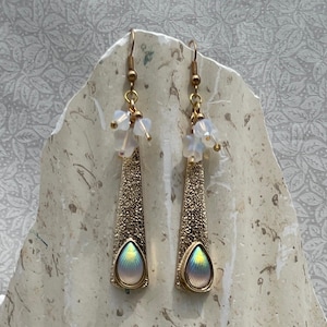 May include: A pair of gold-toned earrings with a textured, elongated design. Each earring features a teardrop-shaped iridescent center and is adorned with small, clear beads. The earrings hang from gold-colored hooks.