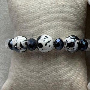 May include: A beaded bracelet featuring three large, round, white beads with black speckled patterns, alternating with smaller, faceted, dark metallic-colored beads. The beads are strung together in a linear arrangement on a clear elastic cord.