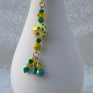 May include: A gold bookmark with a yellow and green glass bead charm. The charm has a polka dot pattern and is attached to a chain with smaller green and yellow beads.