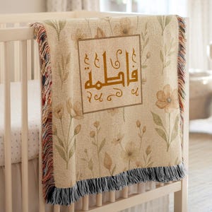 May include: A beige baby blanket with a floral pattern and the name "Fatima" in gold script within a square frame. The blanket has a fringed edge with red, black, and beige threads. It is draped over a white crib.