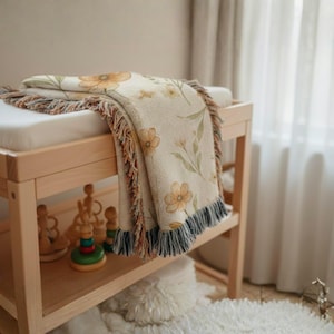 May include: A woven throw blanket featuring a cream-colored base with a repeating pattern of soft orange and yellow flowers and green leaves. The blanket has a textured finish and decorative fringe edges in shades of blue, brown, and cream.