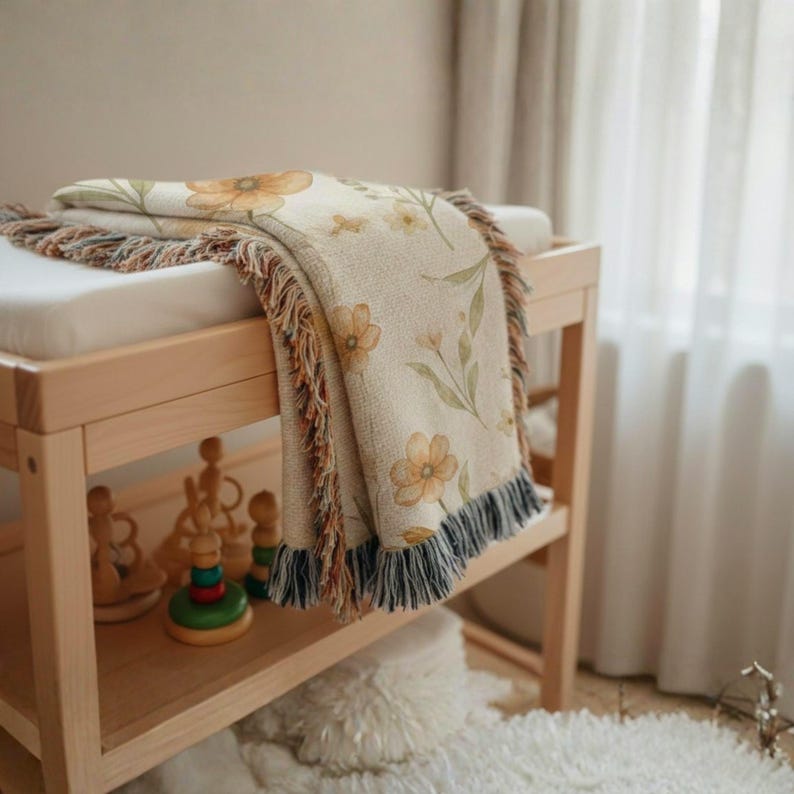May include: A woven throw blanket featuring a cream-colored base with a repeating pattern of soft orange and yellow flowers and green leaves. The blanket has a textured finish and decorative fringe edges in shades of blue, brown, and cream.