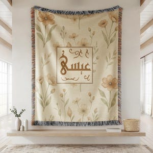 May include: A woven throw blanket featuring a cream-colored background with a repeating pattern of watercolor-style tan flowers and green leaves. In the center, a rectangular frame contains brown Arabic calligraphy with decorative flourishes.