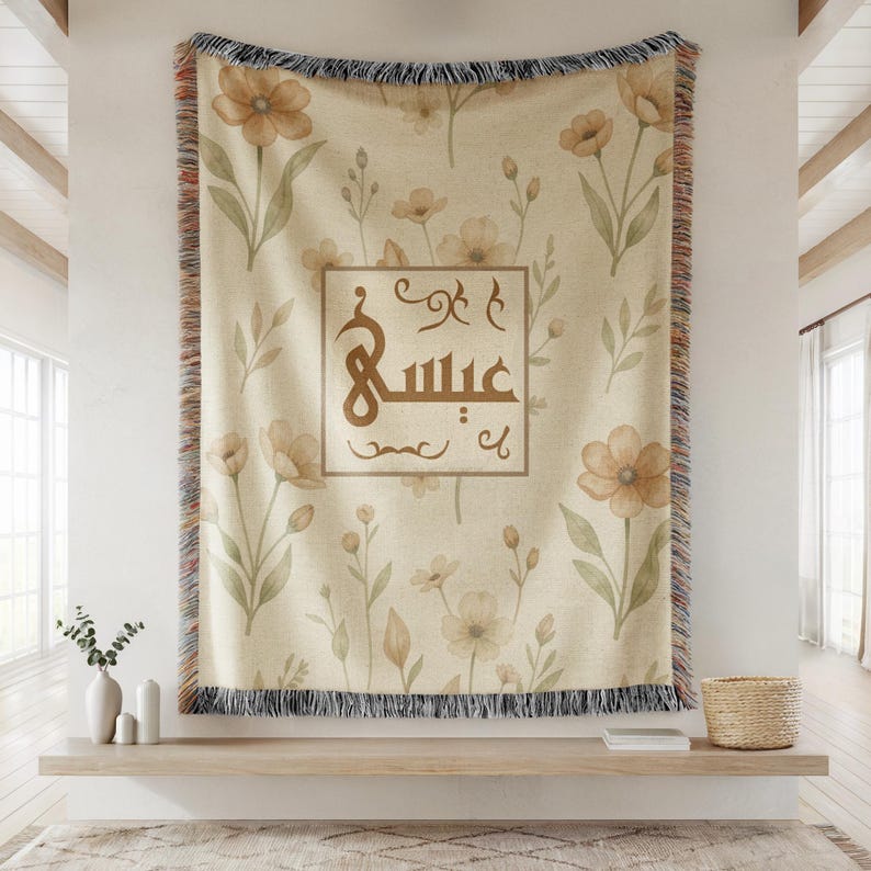 May include: A woven throw blanket featuring a cream-colored background with a repeating pattern of watercolor-style tan flowers and green leaves. In the center, a rectangular frame contains brown Arabic calligraphy with decorative flourishes.