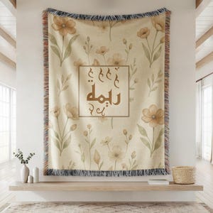 May include: A woven throw blanket featuring a cream background with a repeating pattern of light brown and green watercolor-style flowers. A central rectangular frame contains brown Arabic calligraphy text centered on the fabric with fringed edges.