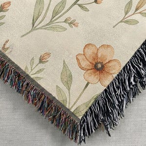 May include: A woven throw blanket featuring a cream-colored background with a botanical pattern of orange flowers and green leaves. The edges of the blanket are finished with a decorative fringe that incorporates multicolored threads and black fibers.