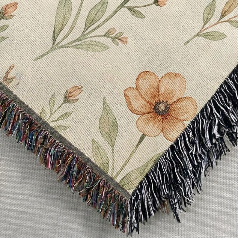 May include: A woven throw blanket featuring a cream-colored background with a botanical pattern of orange flowers and green leaves. The edges of the blanket are finished with a decorative fringe that incorporates multicolored threads and black fibers.