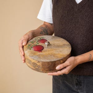 Peut inclure: Une boîte de rangement ronde en bois avec une finition à grain naturel, présentant une illustration peinte à la main de deux grenades rouges sur une branche verte sur le couvercle. Le couvercle inclut un petit bouton métallique argenté.