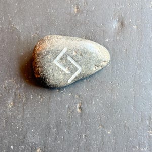 May include: A gray, smooth stone with a white rune symbol etched on its surface. The stone is oval-shaped and appears to be naturally weathered. The rune is a stylized angular design.