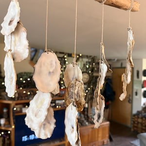 May include: A hanging mobile featuring multiple white and tan oyster shells suspended from a horizontal piece of driftwood by thin twine. The shells are arranged in vertical strands, showcasing their natural iridescent interiors and textured exteriors.