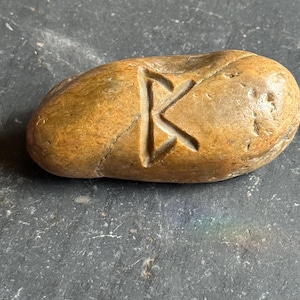 May include: A smooth, oval-shaped stone with a carved letter "K". The stone is a warm brown color with variations in tone. The carved letter is a lighter shade, creating a contrast. The stone appears to be natural and weathered.
