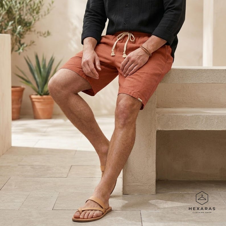 May include: Rust-colored shorts with a drawstring waist and a black long-sleeve shirt. The person is wearing light brown leather sandals. The image is taken outdoors, with a neutral background and a logo in the bottom right corner.